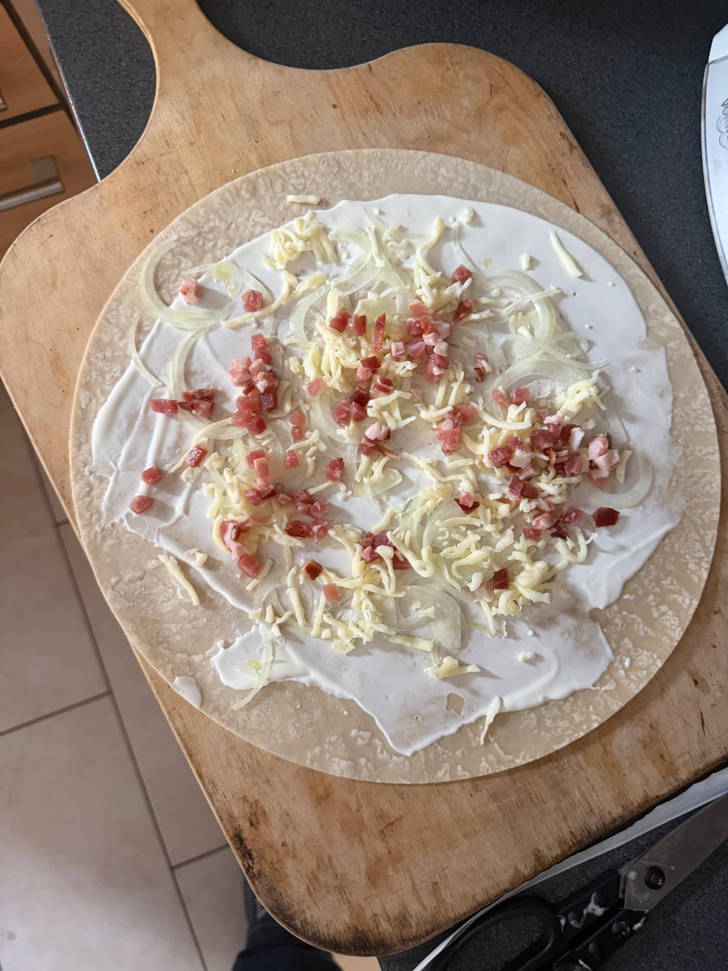 Ungebackener Flammkuchen auf einer Holz-Schaufel, belegt mit Crème fraîche, Zwiebeln, Schweinebäckchen und Mozzarella.
